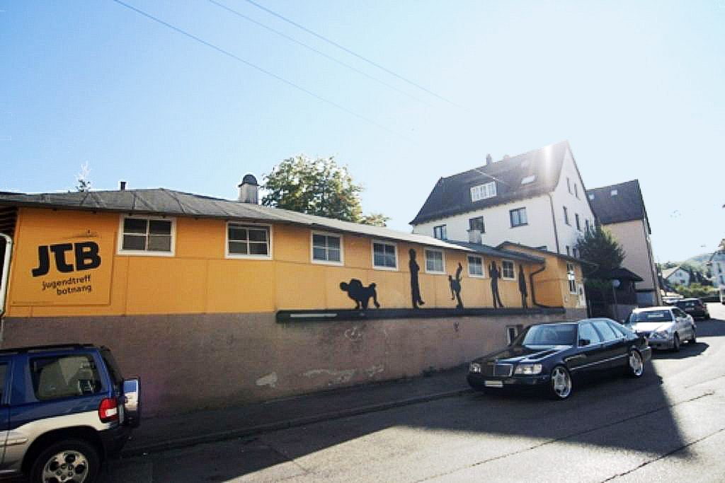 Gelbes Gebäude mit der Aufschrift „JTB Jugendtreff Botnang“ und Silhouetten von Jugendlichen an der Fassade. Autos parken davor.