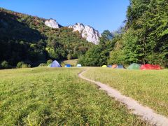 Wiese mit bunten Zelten vor einer bewaldeten Felskulisse unter blauem Himmel.