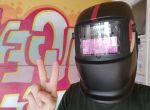 Eine Person trägt einen Schweißhelm und zeigt das Peace-Zeichen, im Hintergrund eine bunte Wand.