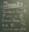 Tafel mit handgeschriebener Snackkarte, u. a. Sandwich, Pizza, Burger, Müsli und Milkshake mit Preisen.