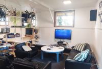 Gemütliche Sitzecke mit schwarzen Ledersesseln und Sofa, kleinem weißen Tisch mit PlayStation-Controllern, Fernseher an der Wand und Regalen voller Spiele und Pflanzen im Hintergrund.