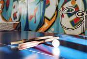Tischtennisschläger und Ball liegen auf einer Tischtennisplatte, im Hintergrund bunte Graffiti-Wand.