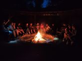 Gruppe Jugendlicher sitzt abends um ein großes Lagerfeuer, das die Szene beleuchtet.